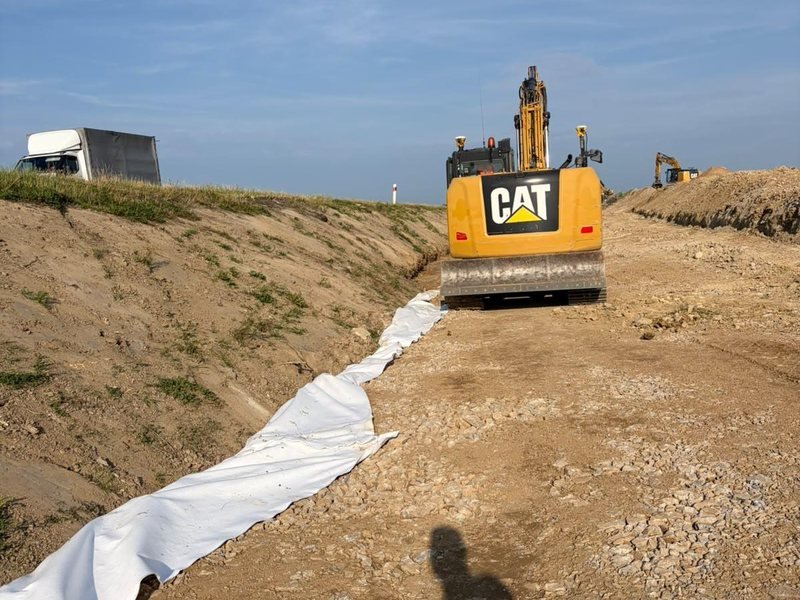 Rypadlo CAT pokládá geotextilii na násypu silnice